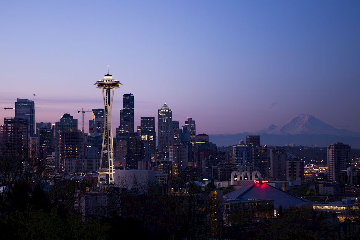 The image shows a city skyline at dusk with a prominent tower, likely the Space Needle, and distant mountains in the background.