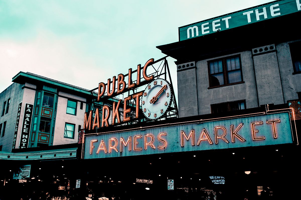 The image shows a neon sign for a public farmers market, with buildings in the background and an overcast sky.