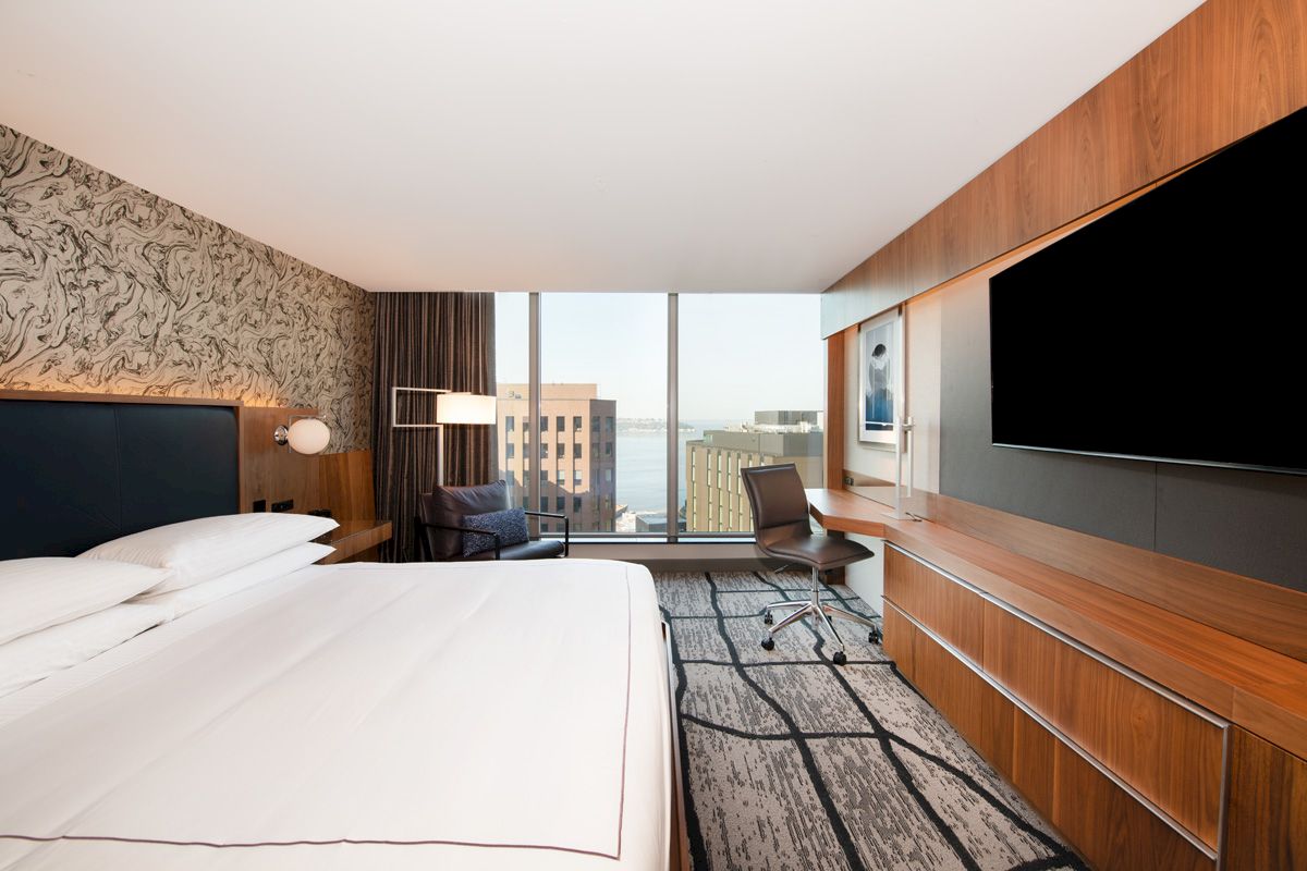 The image shows a modern hotel room featuring a large bed, desk, armchair, and a window with a city view.