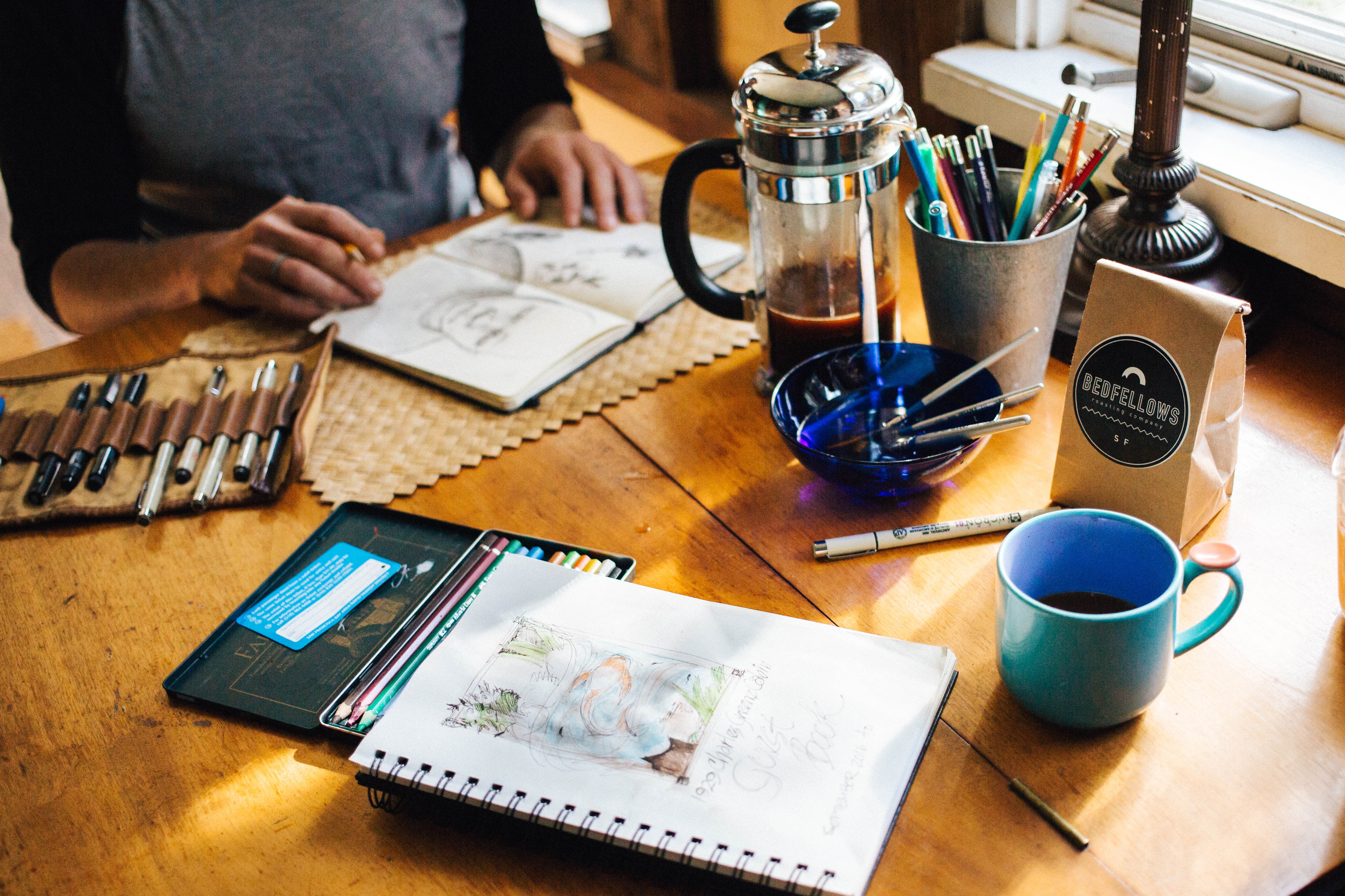 The image shows a cozy workspace with sketchbooks, art supplies, coffee, and a person drawing at a wooden table.