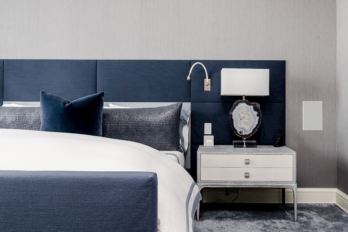 The image features a stylish bedroom with a navy headboard, decorative pillows, a bedside table, and a lamp. The decor is modern.